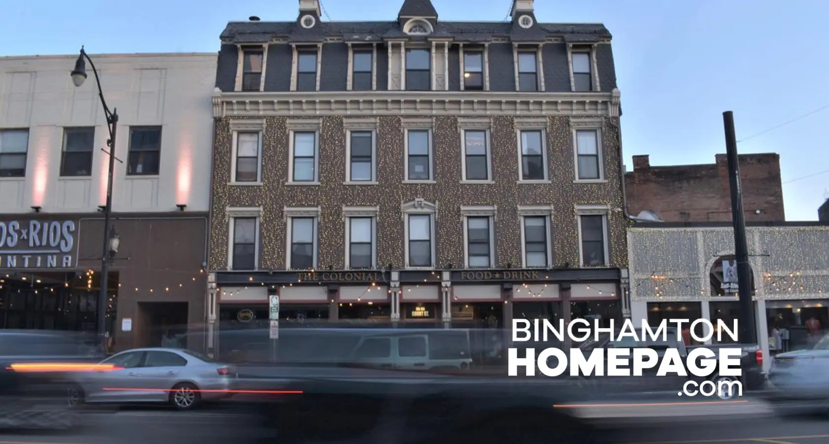 "The Colonial restaurant building in Binghamton decorated with string lights, cars passing by, Binghamton Homepage logo."