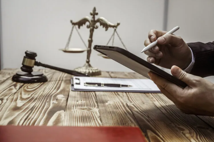 A businessman in a formal suit signs a contract, hands close up, suggesting possible bribery, at a wooden desk with legal documents, scales, and a wooden gavel representing legislation.